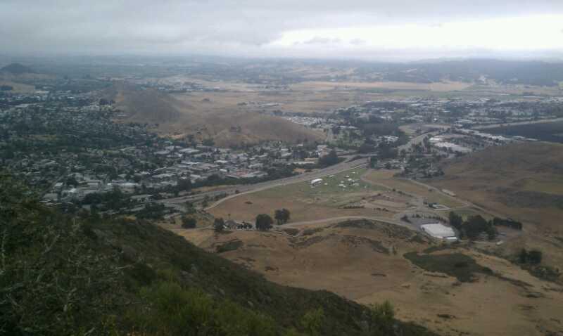 A scenic panoramic view from a mountain overlooking a valley and a small town, with a mixture of urban and rural landscapes. The sky is overcast with gray clouds, and the terrain includes patches of open land and roads winding through the area. Madonna Mountain: Rock Garden mountain bike trail.