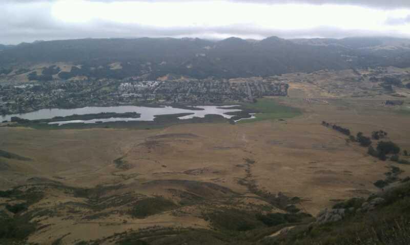 A panoramic view of a landscape featuring a mix of grassy plains and hills, with a small body of water and a residential area visible in the background. The scene is set under a cloudy sky, showcasing a combination of natural and urban environments. Madonna Mountain: Rock Garden mountain bike trail.