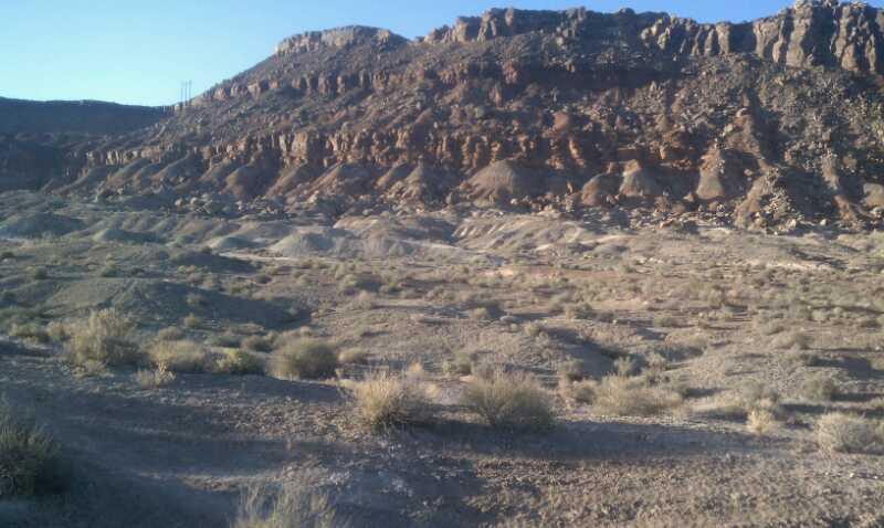 A vast desert landscape featuring a rugged, rocky plateau in the background, with a foreground of sparse vegetation and rolling sandy hills under a clear blue sky. Bearclaw Poppy mountain bike trail.