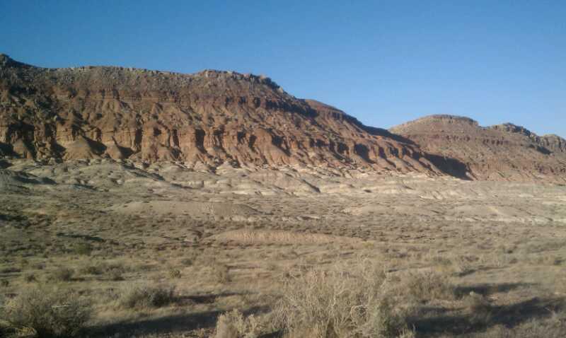 A panoramic view of rugged desert terrain featuring layered sedimentary rock formations against a clear blue sky. The landscape includes dry grasses and shrubs in the foreground, with steep, textured cliffs rising in the background. Bearclaw Poppy mountain bike trail.