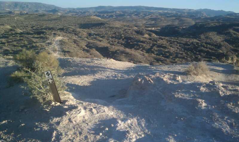 A view of a rocky, arid landscape with rolling hills and distant mountains under a clear blue sky. In the foreground, there's a trail marker indicating a path, surrounded by sparse vegetation. The scene conveys a sense of wilderness and natural beauty. Bearclaw Poppy mountain bike trail.