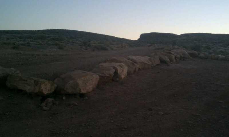 A rocky pathway leading through a desert landscape at dusk, with large stones arranged along the ground and rolling hills in the background. The scene is bathed in soft twilight. Bearclaw Poppy mountain bike trail.