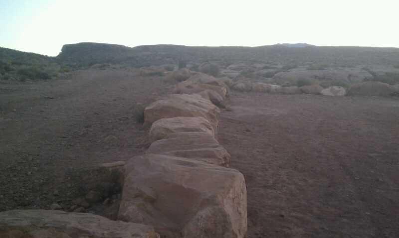 A rocky landscape with a line of large stones marking a path or boundary, set against a backdrop of rolling hills under a clear sky. The scene is arid and natural, suggesting a remote outdoor area. Bearclaw Poppy mountain bike trail.