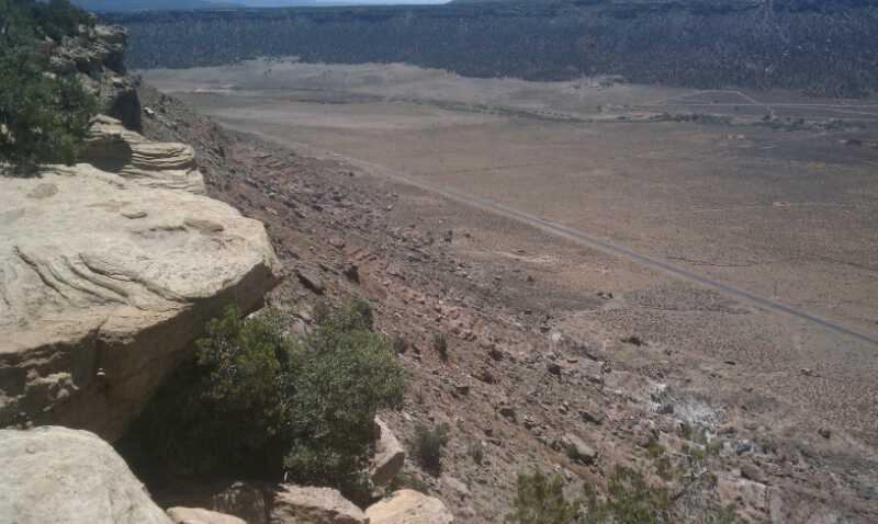 A panoramic view from a rocky cliff edge overlooking a vast desert landscape, featuring layered rock formations and sparse vegetation. In the distance, a winding road cuts through the arid terrain, under a clear blue sky. Gooseberry Mesa mountain bike trail.