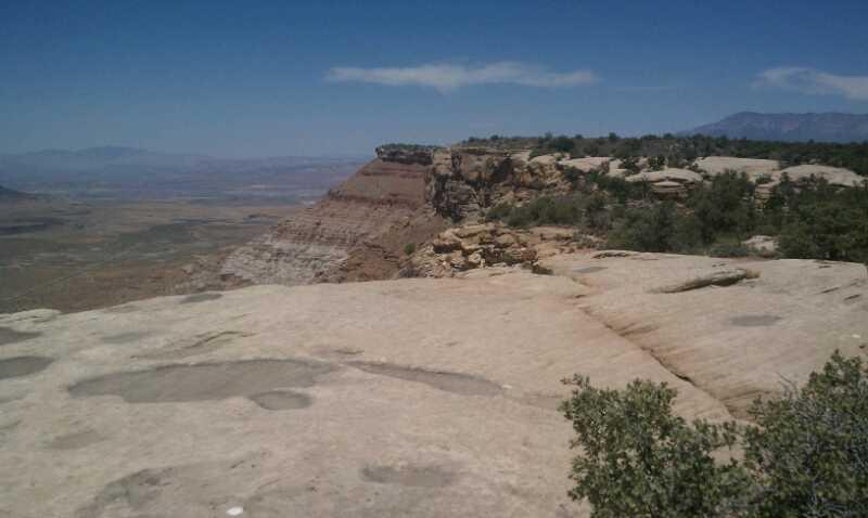 A panoramic view of a rocky cliffside with layered geological formations, showcasing a vast landscape under a clear blue sky. In the foreground, light-colored rocky terrain is visible, while the background features distant mountains and valleys. Sparse vegetation, including small shrubs, is seen along the cliff edge. Gooseberry Mesa mountain bike trail.
