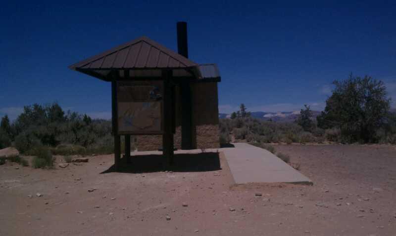 A small, rustic structure with a brown metal roof and a signboard stands in a dusty, open area surrounded by sparse vegetation. In the background, distant mountains can be seen under a clear blue sky. Gooseberry Mesa mountain bike trail.