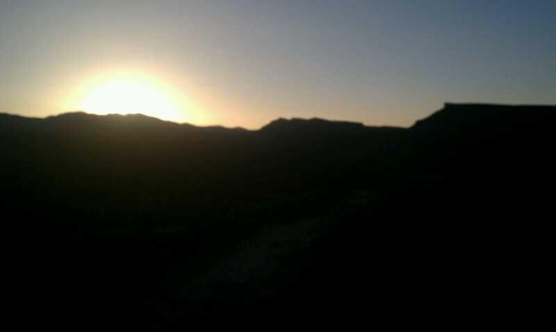 Silhouette of mountains against a sunset sky, with the sun peeking over the horizon, casting a warm glow. Gooseberry Mesa mountain bike trail.