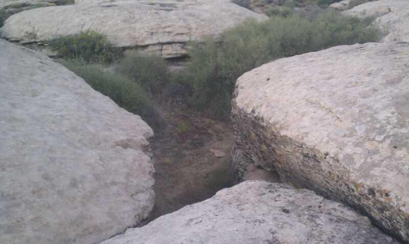 A rocky landscape featuring large, flat boulders with sparse vegetation in the background. A narrow, sandy pathway can be seen winding between the rocks, suggesting a natural divide in the terrain. Zen Trail mountain bike trail.