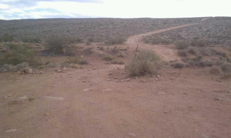 A dry, rugged landscape with a dirt path winding through sparse vegetation. In the foreground, there are scattered rocks and a few bushes, while the background features rolling hills under a cloudy sky. The scene conveys a remote, arid environment. Zen Trail mountain bike trail.