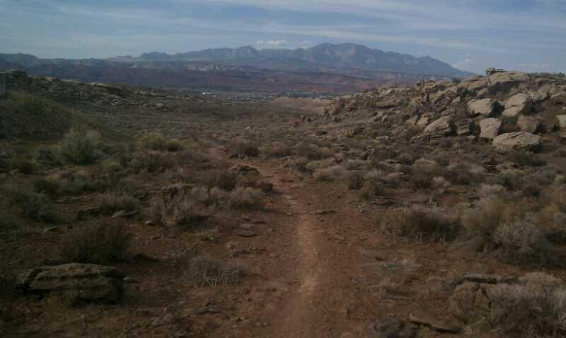 A dirt trail winding through a rocky, arid landscape with sparse vegetation, leading toward distant mountains under a partly cloudy sky. Barrel Roll mountain bike trail.