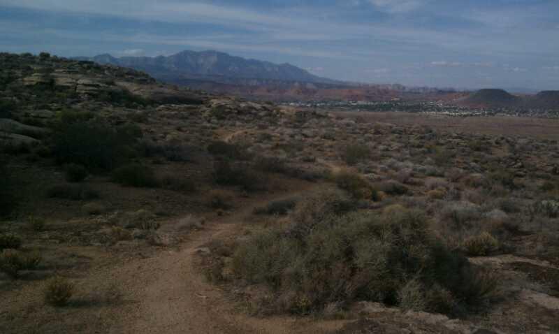 A natural landscape featuring a winding dirt path leading through shrubs and grasses, with rocky hills in the foreground and a range of mountains visible in the background under a partly cloudy sky. The scene captures the essence of an open, arid environment. Barrel Roll mountain bike trail.