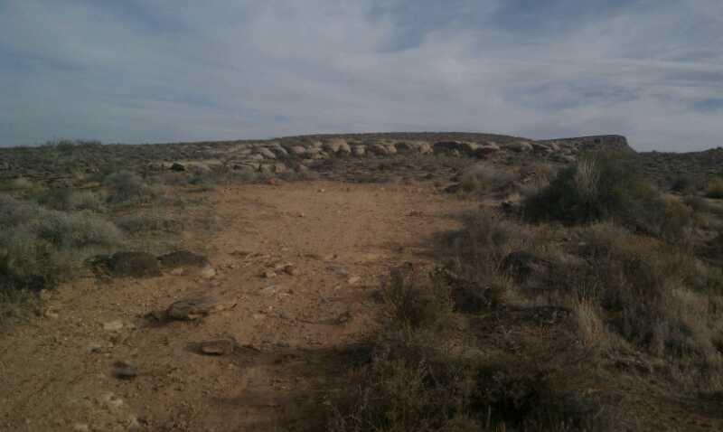 A dirt path leading through arid terrain, surrounded by sparse vegetation and low shrubs, with a distant rocky formation under a cloudy sky. Barrel Roll mountain bike trail.