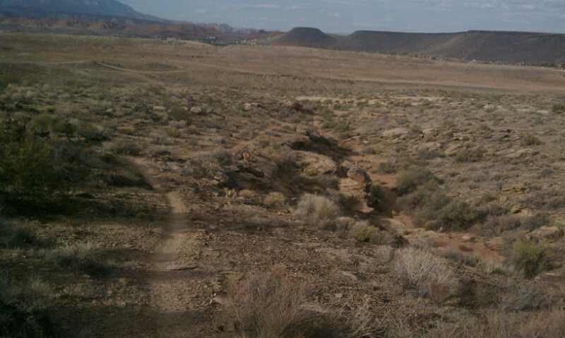 A sandy, arid landscape with sparse vegetation and a winding dirt path leading through the terrain. The background features rocky hills and an expansive sky with scattered clouds. Barrel Roll mountain bike trail.