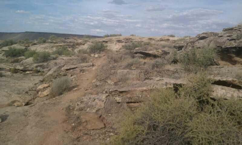 A rugged landscape featuring rocky terrain with a dirt path winding through sparse vegetation. The sky is partly cloudy, and distant hills are visible in the background. Barrel Roll mountain bike trail.