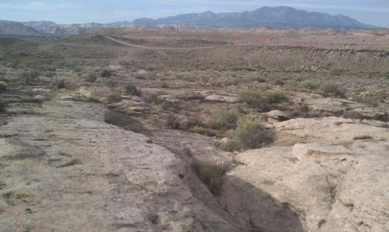 A vast, rocky landscape with scattered shrubs and vegetation, featuring distant mountains under a partly cloudy sky. The foreground shows a rocky terrain with a slight incline leading to the horizon, where the mountainous backdrop is visible. Barrel Roll mountain bike trail.