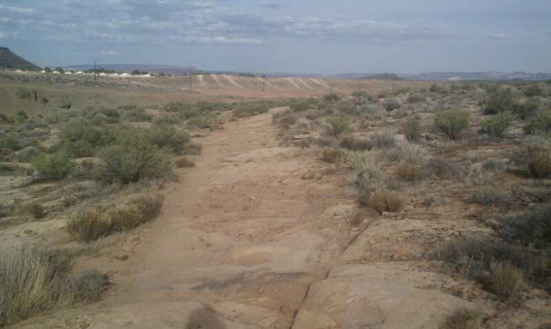 A wide, sandy path stretches through a desert landscape, surrounded by sparse bushes and low vegetation. In the background, a distant hilly terrain and a cloudy sky are visible, with a road or pathway lined by power poles. The scene conveys a sense of arid wilderness. Barrel Roll mountain bike trail.