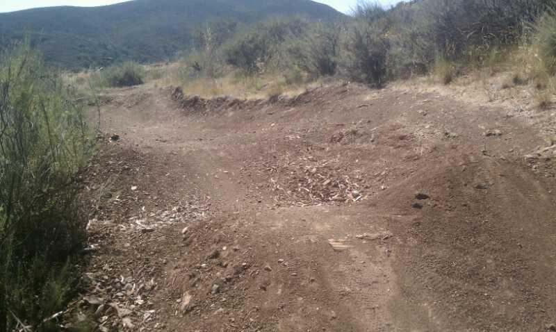 A dirt path winding through a natural landscape with sparse vegetation and hills in the background. The ground is uneven with a slight rise, indicating it may be a trail used for hiking or biking. The Eucs Project / Elevator mountain bike trail.