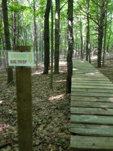 A wooden pathway leading through a forest, with a caution sign reading "Caution: Big Drop" placed beside it. The area is surrounded by tall trees and lush greenery, with fallen leaves covering the ground. Anderson Park mountain bike trail.