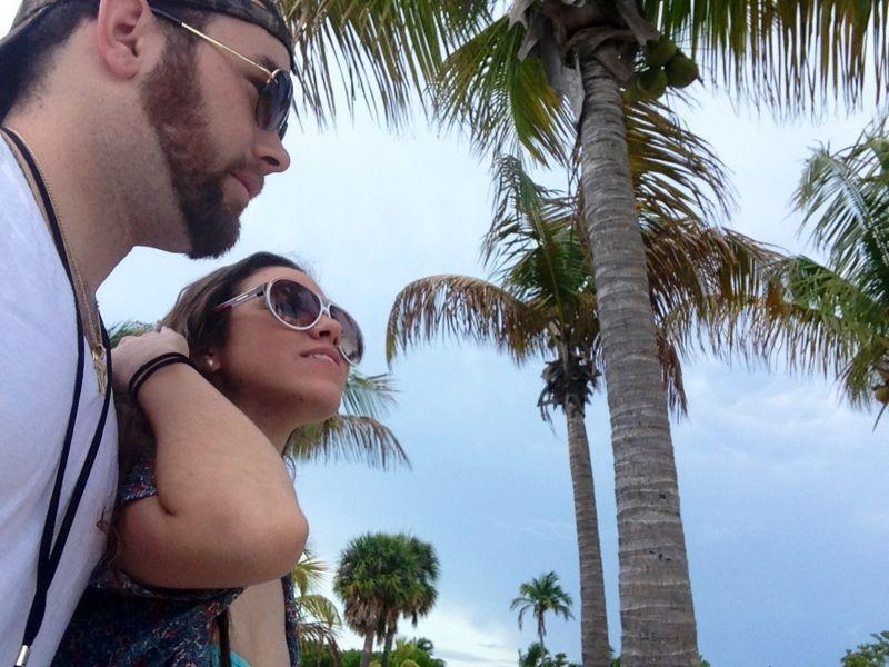 A couple walking side by side in a tropical setting, surrounded by palm trees. The man wears sunglasses and a casual T-shirt, while the woman also sports sunglasses and a colorful top. The background features a cloudy sky, adding a relaxed, beach-like atmosphere. North Port Mountain Bike Trails mountain bike trail.
