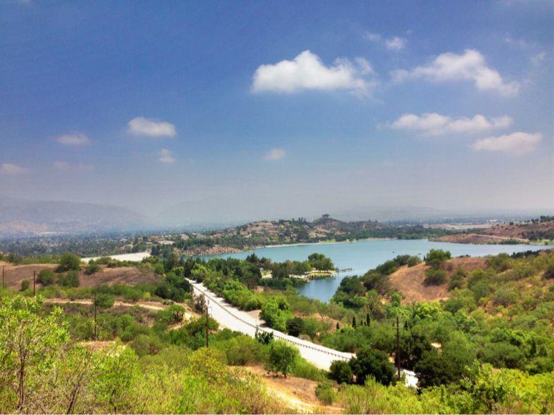 A panoramic view of a serene landscape featuring a winding road alongside a lake, surrounded by lush greenery and rolling hills under a partly cloudy sky. Frank G. Bonelli Regional Park mountain bike trail.
