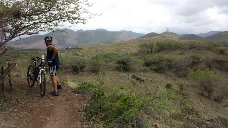 A cyclist standing next to a mountain bike on a dirt trail, surrounded by sparse vegetation and rolling hills under a cloudy sky. The cyclist is wearing a helmet and athletic clothing, with mountain ranges visible in the background. Los Pinchos mountain bike trail.