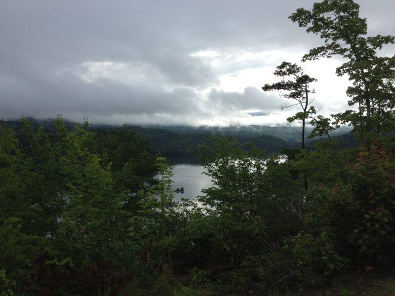 A serene landscape featuring a calm lake surrounded by lush green trees and rolling hills, with a cloudy sky overhead that creates a misty atmosphere. Tsali Mouse Branch Loop mountain bike trail.