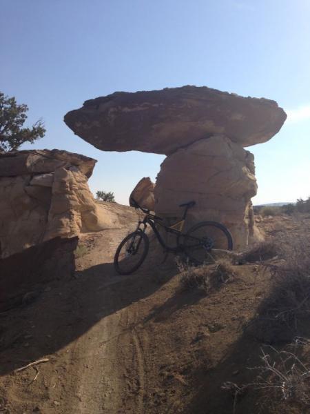 A mountain bike rests against a large rock formation resembling a mushroom, set against a clear sky. The landscape features sandy terrain and sparse vegetation, showcasing the natural beauty of the area. High Desert Trail System mountain bike trail.