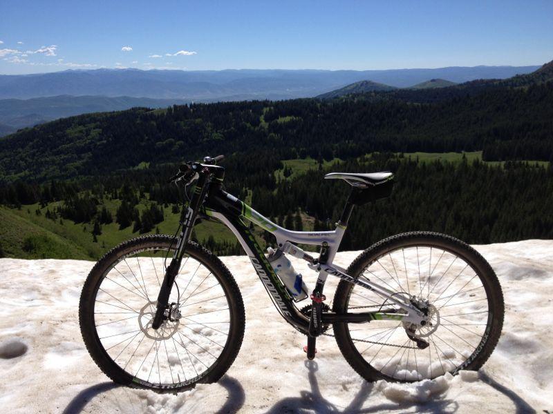 A mountain bike positioned on a patch of snow, overlooking a scenic view of green hills and mountains under a clear blue sky. Wasatch Crest mountain bike trail.