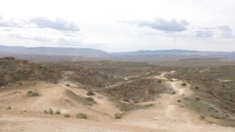 A vast, arid landscape featuring rolling hills and a rocky terrain. The scene is set under a cloudy sky, with distant mountains visible on the horizon. Sparse vegetation dots the sandy ground, and winding trails can be seen traversing the terrain. Bearclaw Poppy mountain bike trail.
