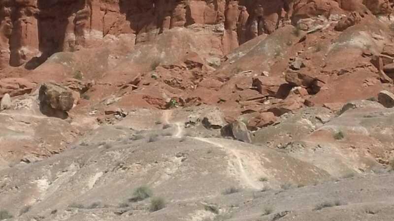 A wide view of a rugged, rocky landscape featuring red-hued cliffs and a dirt path winding through the barren terrain. The scene captures a natural area with patches of sparse vegetation and large boulders scattered throughout. Bearclaw Poppy mountain bike trail.