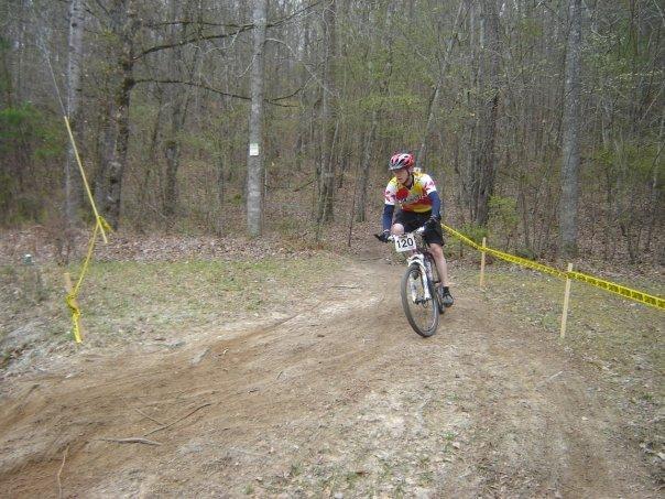 A cyclist wearing a colorful jersey and helmet rides a mountain bike along a dirt trail in a wooded area. Yellow caution tape is visible on either side of the path, indicating a race course. Trees in the background show early signs of spring, with a mix of bare branches and budding greenery. The cyclist's number is visible as "120." Arrowhead Park mountain bike trail.