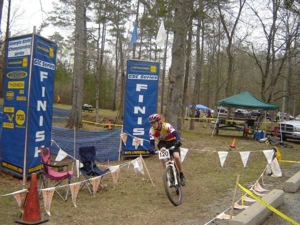 A cyclist in a red and yellow jersey competes in a mountain biking event, crossing the finish line marked by large banners that say "FINISH." The scene is set in a wooded area with trees in the background, and there are camping chairs and tents visible, indicating a race event atmosphere. Arrowhead Park mountain bike trail.