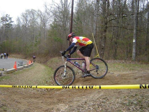 A cyclist in a red, yellow, and black racing jersey is navigating a dirt path on a mountain bike. The rider is airborne, with the front wheel lifted off the ground, while the rear wheel remains planted. Yellow caution tape is visible on the ground, and orange cones are positioned nearby, indicating a racing event. The background features a wooded area with sparse trees and no leaves, suggesting an early spring setting. Arrowhead Park mountain bike trail.