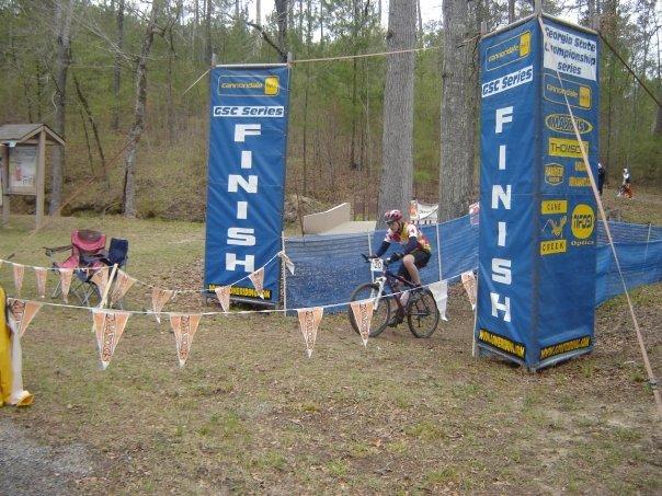 A young cyclist approaches the finish line marked by large blue banners that read "FINISH," with a garland of colorful flags hanging nearby. The scene is set in a wooded area, with trees in the background and a couple of bicycles resting on the ground. Arrowhead Park mountain bike trail.