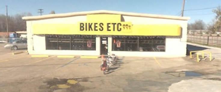 A storefront of a bike shop named "Bikes Etc." featuring a bright yellow awning and large windows displaying bicycles. The shop is surrounded by a parking lot with a few bikes parked outside. The clear blue sky adds to the scene.