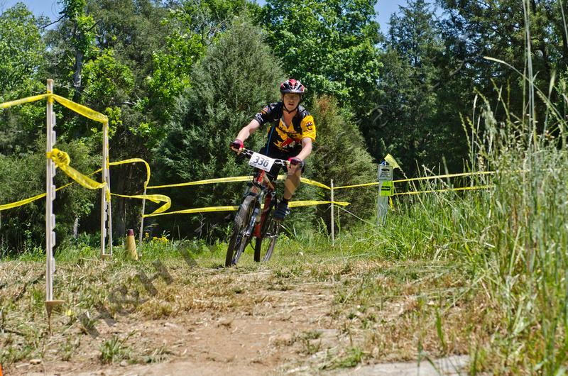A cyclist in a black and yellow jersey rides a mountain bike along a dirt path marked by yellow caution tape in a wooded area. The cyclist is focused and wearing a helmet, with a race number 396 displayed on their bike. Lush greenery surrounds the track under a clear blue sky. Georgia International Horse Park mountain bike trail.