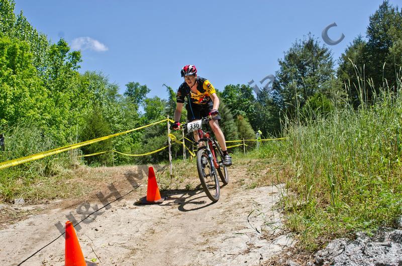 A mountain biker is riding along a dirt path in a wooded area, smiling as they navigate the trail. They are wearing a yellow and black cycling jersey and a helmet, with a race number, 336, visible on their bike. Orange cones are placed along the path, and a yellow tape marks the boundaries of the trail. Lush greenery surrounds the area under a clear blue sky. Georgia International Horse Park mountain bike trail.