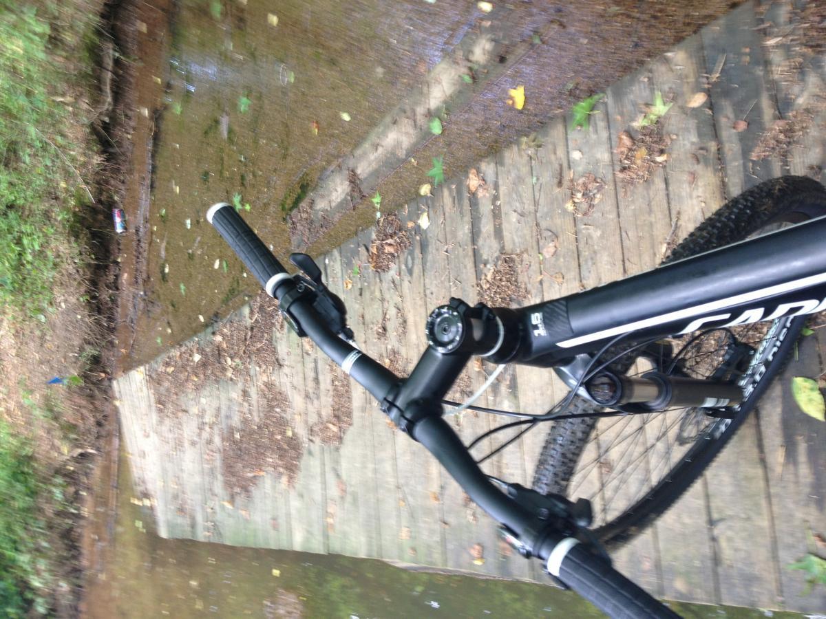 Image of a mountain bike resting on a wooden path beside a shallow stream. The handlebars are visible in the foreground, with the path covered in leaves and scattered debris. The stream reflects greenery from the surrounding environment. Charleston Park mountain bike trail.
