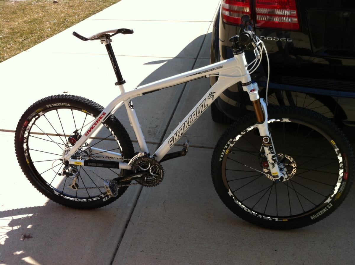 Santa Cruz Chameleon: A white mountain bike with a sleek design and knobby tires parked on a concrete driveway next to a black vehicle. The bike features notable components such as a suspension fork, disc brakes, and a prominent brand logo on the frame. Sunlight casts shadows, highlighting the bike's details and the surrounding grass.