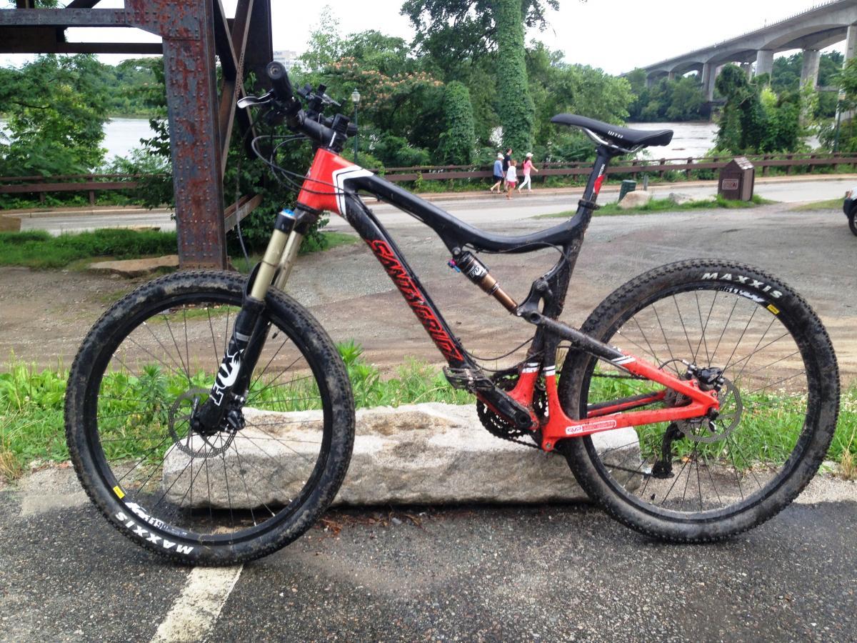 Santa Cruz Blur TRC: A mountain bike with a black and red frame is parked on a stone near a path, surrounded by greenery. In the background, there is a river and a bridge, with people walking along the path. The bike has dirt on its tires, indicating it has been used off-road.