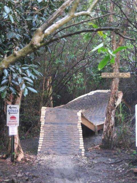 A wooden pathway leading through a dense forest, with a signpost labeled "Pipeline" directing the way. On the left, a warning sign states "BYPASS" with additional cautionary messages. The pathway is slightly elevated and constructed from interlocking wooden planks, surrounded by lush green foliage. Markham Park mountain bike trail.