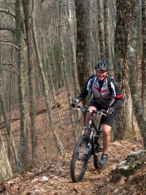 A man riding a mountain bike on a forest trail, surrounded by bare trees. He is wearing a helmet and biking gear, smiling as he navigates the path. The ground is covered in leaves, indicating an autumn or early winter setting. Aska Trail System mountain bike trail.