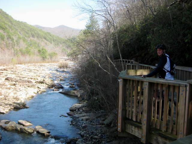 A person in biking gear stands on a wooden overlook railing next to a river with smooth rocks and flowing water. Verdant hills rise in the background under a clear blue sky. The setting conveys a serene outdoor landscape, ideal for nature enthusiasts and cyclists. Rhododendron mountain bike trail.