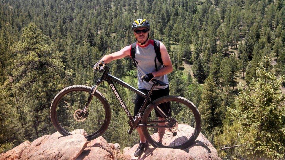 Diamondback Overdrive Comp: A mountain biker stands confidently on a rocky outcrop, holding a black Diamondback mountain bike. He is wearing a helmet, sunglasses, and a sleeveless shirt, with a dense forest of pine trees and a valley visible in the background under a clear blue sky.