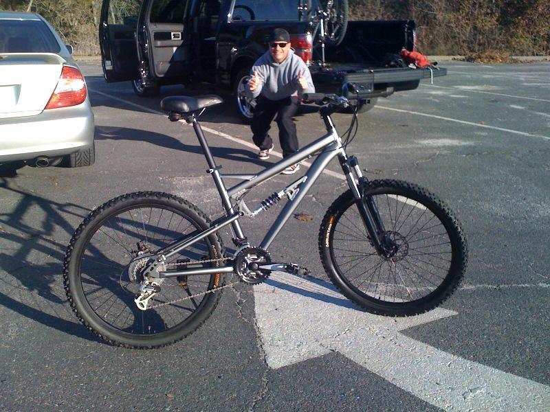 A silver mountain bike is positioned in the foreground, showcasing its thick tires and suspension system. In the background, a person is seen giving a thumbs-up while standing near an open truck with bikes in the back. Several vehicles are parked in a lot surrounded by trees. Granite Bay Trail mountain bike trail.