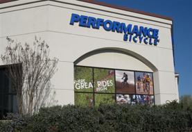 Exterior view of a Performance Bicycle store featuring a prominent blue sign above the entrance and promotional graphics on the windows showcasing cycling activities.