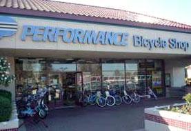 A storefront of a bicycle shop named "Performance Bicycle Shop," featuring bicycles displayed outside. The building has large glass windows and a sign with the shop