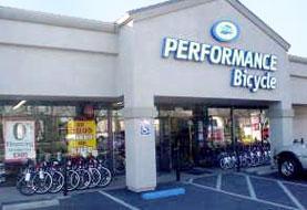 Storefront of a Performance Bicycle shop featuring bicycles displayed outside and large signage on the building. The entrance is visible, along with promotional signs in the windows.
