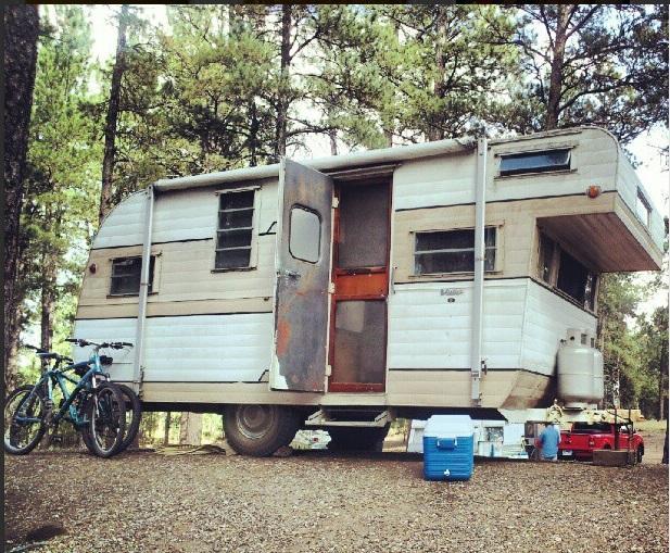 A vintage camping trailer parked in a wooded area, featuring a partially open door and a rustic design. Two bicycles are leaning against the trailer, and coolers sit on the ground nearby. Sunlight filters through the trees, creating a peaceful outdoor setting. Centennial Trail mountain bike trail.