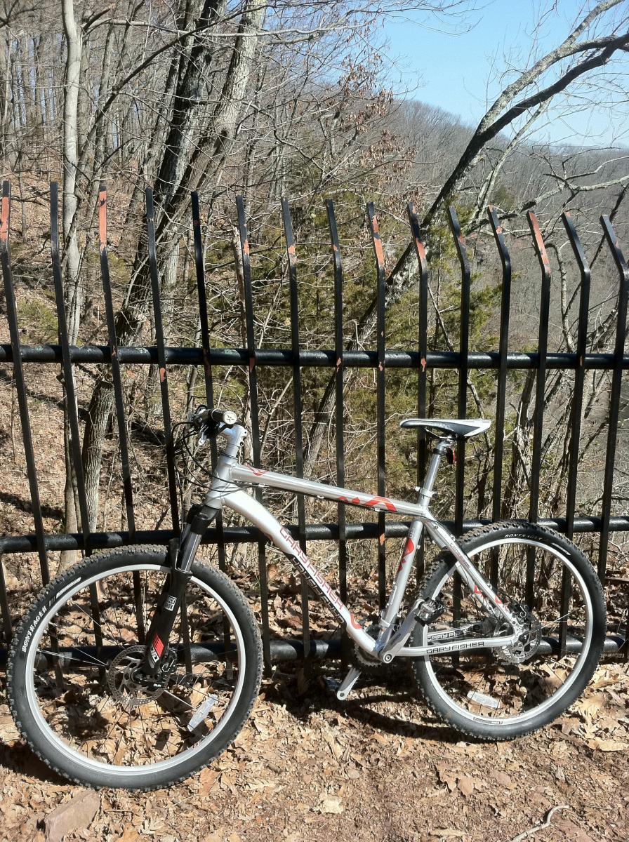 Gary Fisher Piranha: A mountain bike parked against a black wrought iron fence, overlooking a wooded area with bare trees and a clear blue sky. Leaves cover the ground, indicating early spring or late fall.
